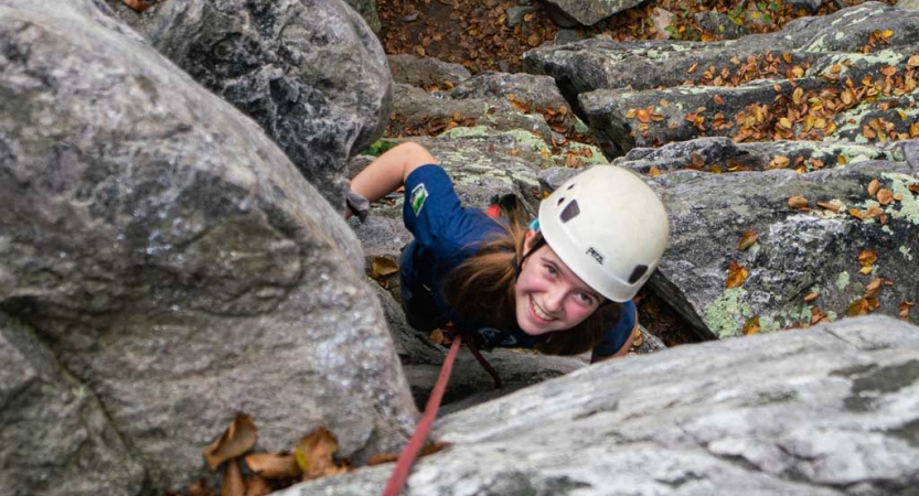 a girl looks up at the camera and smiles while rock climbing on an outward bound course